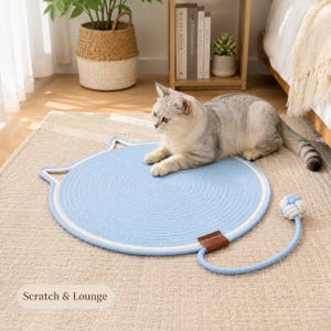 Puede incluir: Una alfombrilla redonda de color azul claro para rascar y descansar, con un borde blanco y una parte superior con forma de orejas de gato. Una cuerda azul claro se extiende desde el lateral y termina en una pequeña pelota de juguete tejida.