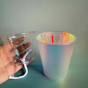 May include: A clear plastic cup with a white cotton wick attached to the base and a separate frosted iridescent cup that displays a rainbow-like sheen on its surface. Both cups feature a tapered cylindrical shape with a wide rim at the top.