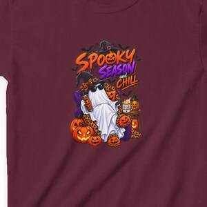 May include: A maroon short-sleeved t-shirt featuring a graphic of a ghost wearing sunglasses and a witch hat, sitting in a chair surrounded by jack-o'-lanterns. The text reads "SPOOKY SEASON and CHILL" in orange and purple lettering with bats.