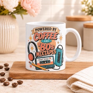以下が含まれることがあります： 「POWERED BY COFFEE AND 80s MIXTAPES」という文字が描かれたカラフルなグラフィック付きの白いセラミック製マグカップ。黒いヘッドフォン、青いポータブルカセットプレーヤー、「Good Songs」と書かれたカセットテープ、黄色い鉛筆のイラストがデザインされています。