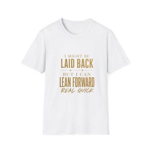 Peut inclure: Un t-shirt blanc à manches courtes avec un texte doré sur la poitrine qui indique : I MIGHT BE LAID BACK + + BUT I CAN LEAN FORWARD REAL QUICK. Le t-shirt présente une encolure ronde et une coupe standard, avec le motif centré sur le devant.