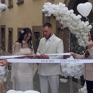 Könnte beinhalten: Eine Hochzeitszeremonie mit einer Person in einem perlenbesetzten weißen Kleid und einer Person in einem hellbeigen Anzug, die ein weißes Band mit der Aufschrift MR & MRS ÖZKOYUNCU durchschneiden. Weiße Blumenarrangements und Luftballons.