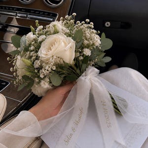 Könnte beinhalten: Ein Blumenstrauß aus weißen Rosen, Schleierkraut und grünen Eukalyptusblättern. Um die Stiele ist ein transparentes weißes Band gebunden, das mit dem Text Selin & Mustafa und dem Datum 24.10.2025 in goldener Schreibschrift bedruckt ist.