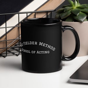 May include: A black ceramic mug with the text "THE FIELDER METHOD SCHOOL OF ACTING" in white. The mug is on a white surface, next to a laptop, a succulent plant in a beige pot, and a wire basket containing notebooks.