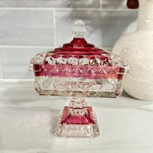 May include: A square glass sweet dish with a lid, featuring a clear and cranberry red diamond-cut pattern. The dish stands on a pedestal base with a matching square foot, and the lid is topped with a small, clear, faceted finial handle.