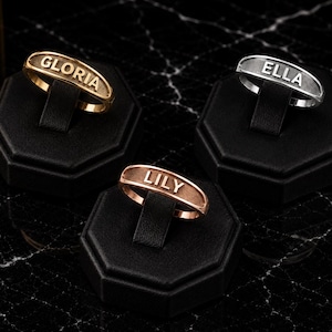 May include: Three metal rings featuring horizontal rectangular plates with engraved names. One gold-toned ring displays the name GLORIA, one silver-toned ring displays the name ELLA, and one rose gold-toned ring displays the name LILY.