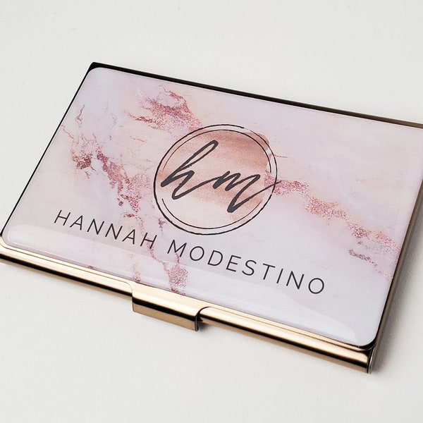 May include: A gold-trimmed business card holder with a pink and gold marble design. The holder features a circle with the initials "HM" in cursive script and the name "HANNAH MODESTINO" printed below.