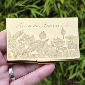 May include: A gold-colored business card holder with the name "Amanda Hammond" engraved on the top. The front of the holder features an engraved floral design of lotus flowers and leaves. The holder is rectangular with rounded edges.