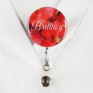 Puede incluir: Un portagafete con un dise&ntilde;o abstracto rojo y dorado, con el nombre "Brittney" impreso en blanco.