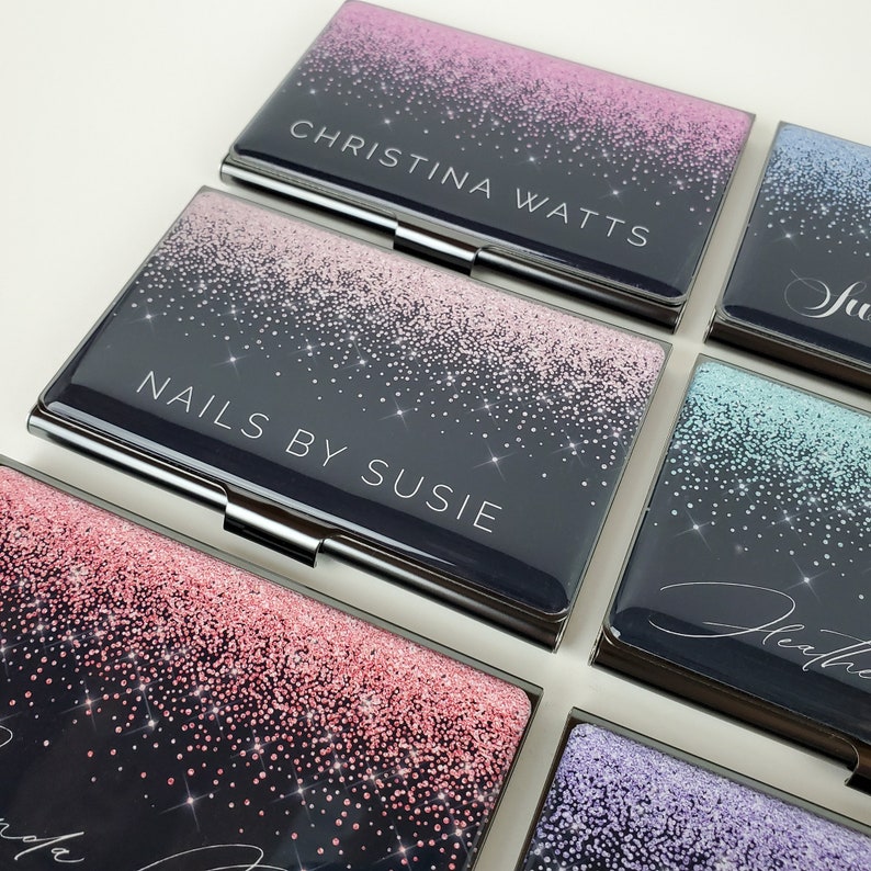 K&ouml;nnte beinhalten: Sechs verschiedene Visitenkartenhalter mit Glitzer-Design. Die Kartenhalter sind schwarz mit einem Farbverlauf aus rosa, blau, gr&uuml;n und lila Glitzer. Der Text auf den Kartenhaltern lautet "Christina Watts", "Nails by Susie", "Julie", "Heather", "Brenda" und "Ashley".