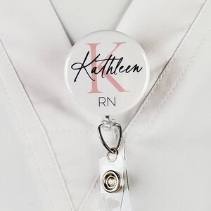 May include: White round badge holder with a pink letter K and the name Kathleen written in black cursive. The badge holder also says RN in black text.