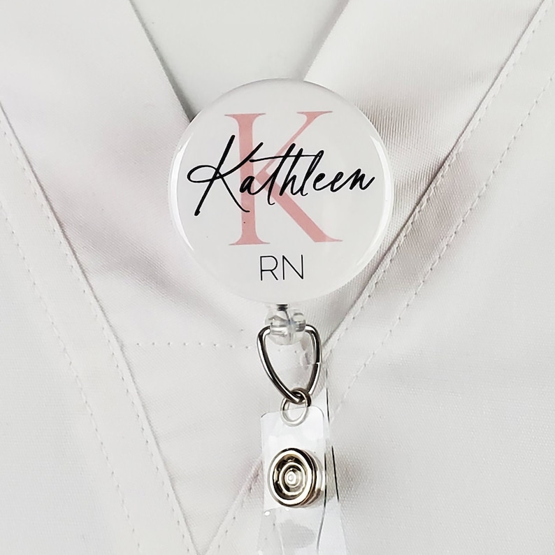 May include: White round badge holder with a pink letter K and the name Kathleen written in black cursive. The badge holder also says RN in black text.