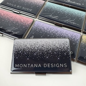 K&ouml;nnte beinhalten: Ein Visitenkartenhalter mit silbernem Glitzerdesign auf schwarzem Hintergrund. Der Text "MONTANA DESIGNS" ist in Wei&szlig; gedruckt.
