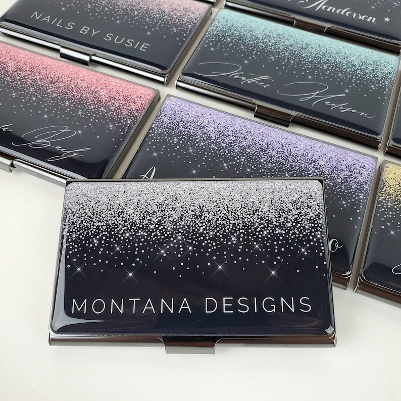 K&ouml;nnte beinhalten: Ein Visitenkartenhalter mit silbernem Glitzerdesign auf schwarzem Hintergrund. Der Text "MONTANA DESIGNS" ist in Wei&szlig; gedruckt.