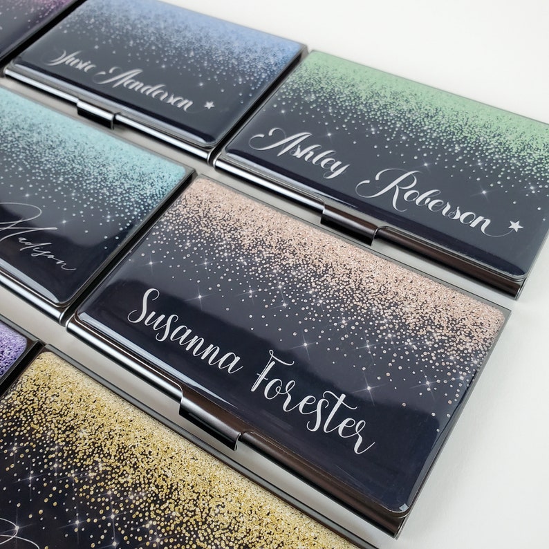 K&ouml;nnte beinhalten: Ein Satz schwarzer Metall-Visitenkartenhalter mit glitzerndem Design. Jeder Halter hat einen anderen Glitzerfarbton und einen in Wei&szlig; gedruckten Namen. Die Namen sind "Susie Anderson", "Ashley Roberson", "Madison", "Susanna Forester".