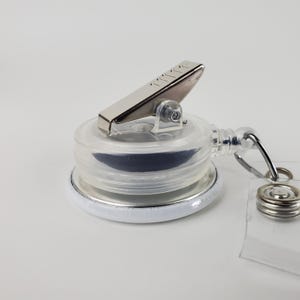 Puede incluir: Un portagafete retr&aacute;ctil blanco con un clip plateado y una caja de pl&aacute;stico transparente. El portagafete tiene una correa de pl&aacute;stico transparente con un anillo plateado.