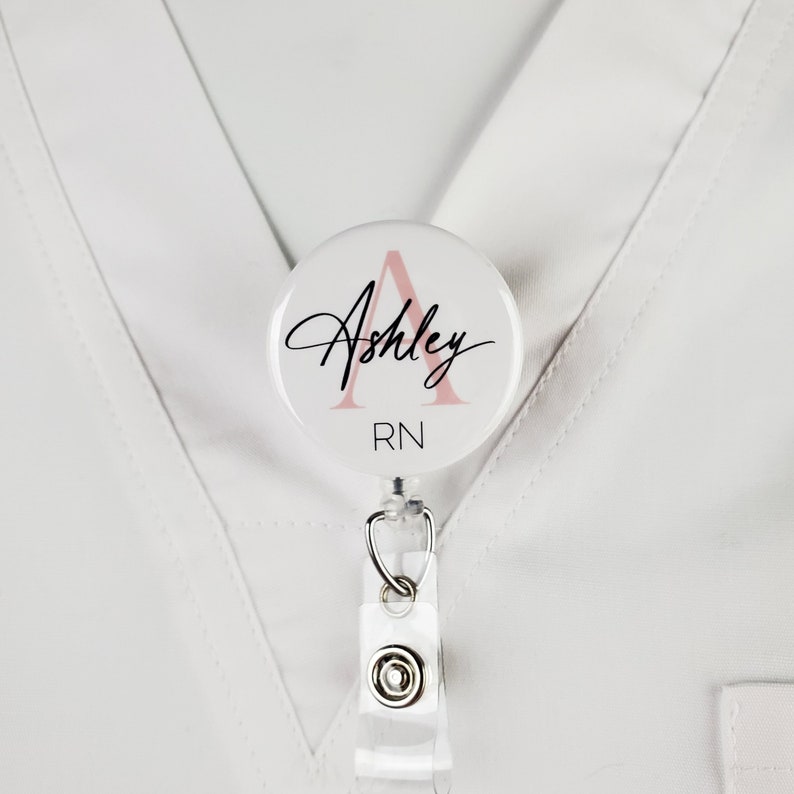 May include: A white badge reel with a pink and white round badge holder. The badge holder has a pink letter A and the text "Ashley RN" in black.