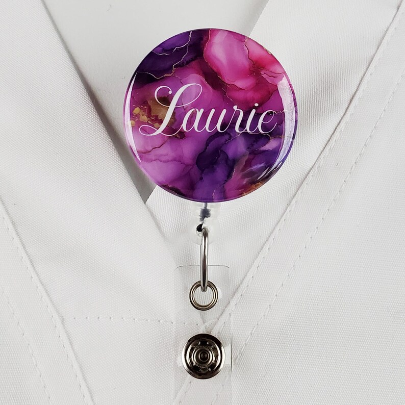 Puede incluir: Un carrete de identificaci&oacute;n con un dise&ntilde;o abstracto en tonos morados y dorados, con el nombre "Laurie" impreso en blanco.