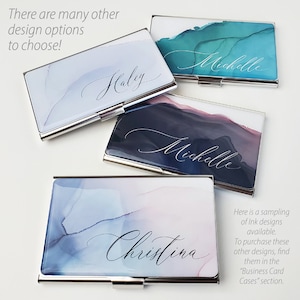 May include: Four silver business card holders with different watercolour designs and names printed on them. The designs include blue and green, blue and pink, and black and grey. The names are Haley, Michelle, Michelle, and Christina.