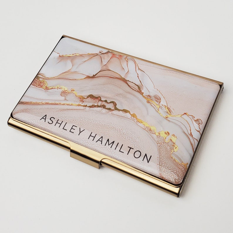 May include: Gold-toned metal business card holder with a pink and gold marble design. The name "ASHLEY HAMILTON" is printed in black on the front.