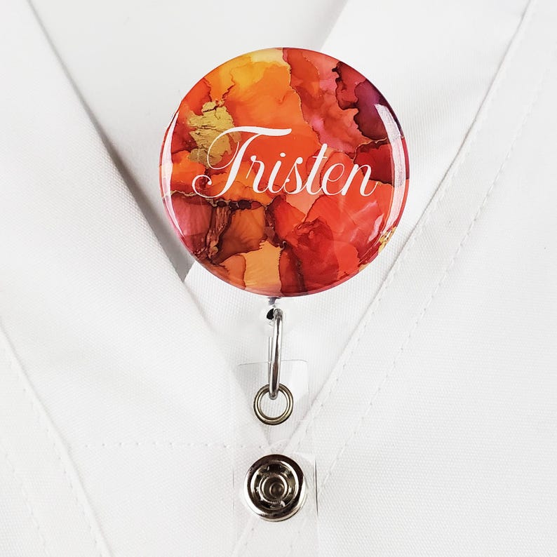 Puede incluir: Un portagafete redondo con un dise&ntilde;o abstracto rojo, naranja y dorado. El nombre "Tristen" est&aacute; impreso en blanco en el portagafete.