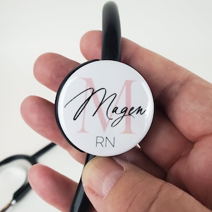 May include: A white stethoscope charm with a pink letter M and the name "Magen" written in black cursive. The charm also has the letters "RN" written in black below the name.
