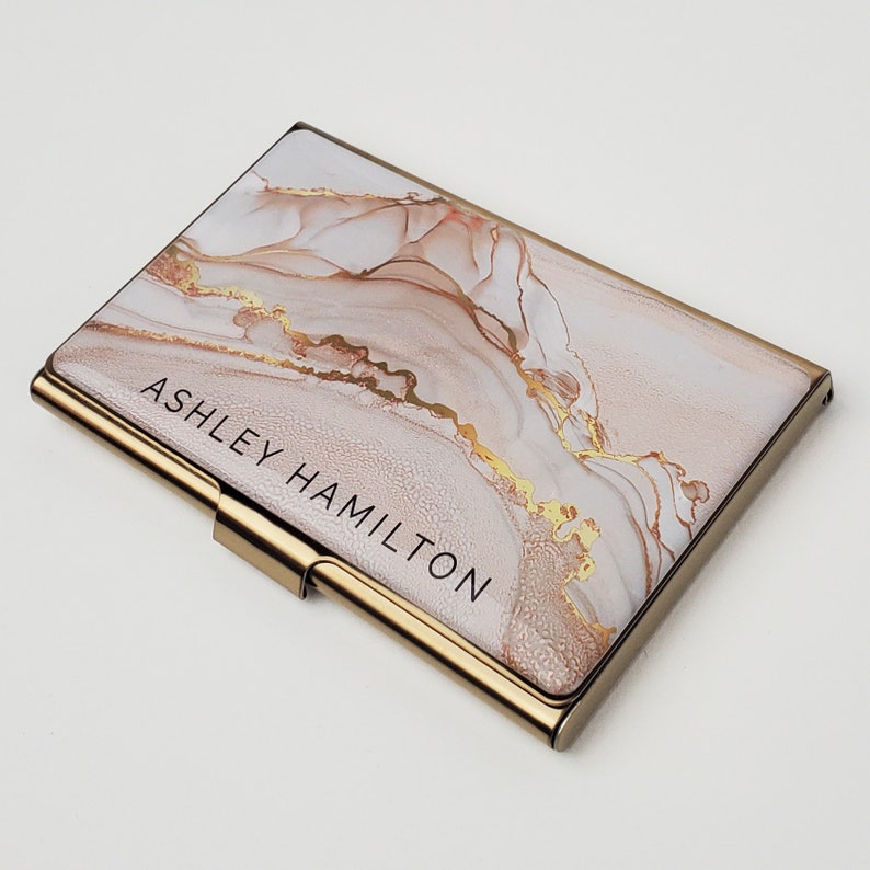 May include: A gold-toned metal business card holder with a white and gold marble design. The holder has the name "ASHLEY HAMILTON" printed on the front.