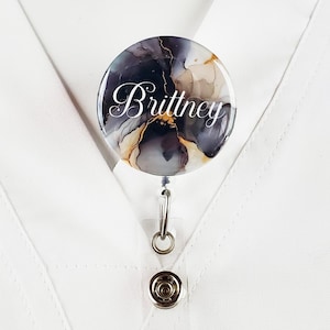 Puede incluir: Un carrete de identificaci&oacute;n con un dise&ntilde;o abstracto negro y dorado, con el nombre "Brittney" impreso en blanco.