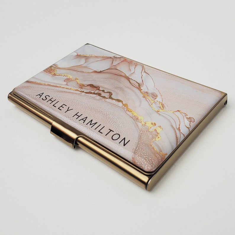 May include: A gold-toned metal business card holder with a pink and white marble design and gold accents. The holder has the name "Ashley Hamilton" printed on the front.