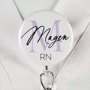May include: White round badge with a purple letter M and the name "Magen" in black script. The badge also has the letters "RN" in black below the name. The badge is attached to a retractable badge reel.