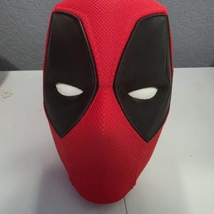 May include: A full-head mask featuring a vibrant red textured mesh fabric exterior. The mask includes two large, almond-shaped black eye patches with white mesh lenses, creating a distinct facial appearance. The design is sleek and form-fitting.