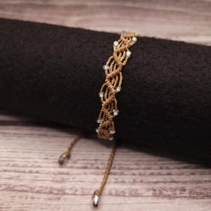 Peut inclure: Un bracelet en macramé beige présentant un motif de cordon tressé complexe agrémenté de petites perles transparentes. Le bracelet comprend deux extrémités de cordon réglables qui pendent, chacune terminée par une petite perle métallique.