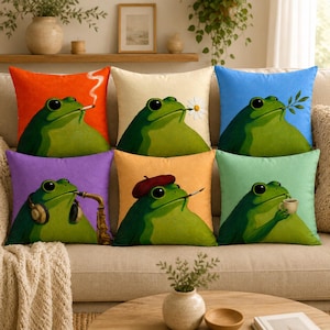 Op de afbeelding: Een set van zes vierkante kussens, elk met een illustratie van een groene kikker tegen een effen achtergrond. De kikkers zijn afgebeeld met verschillende voorwerpen: een sigaret, een wit madeliefje, een groen takje, een koptelefoon met saxofoon, een rode baret met een penseel en een klein kopje koffie.