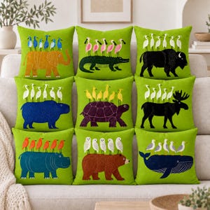 Puede incluir: Un conjunto de nueve cojines cuadrados con un fondo verde brillante. Cada cojín presenta un animal diferente, incluyendo un elefante, cocodrilo, bisonte, hipopótamo, tortuga, alce, oso y ballena, con varias aves posadas sobre sus espaldas.