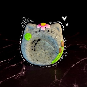 May include: A speckled gray cat-shaped worry stone featuring a pink flower on its head, a green lily pad, and brown cattails. The surface includes small black bird silhouettes and the text Worry Stone written in white script along the side.