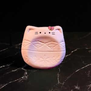 May include: A light pink, cat-shaped ceramic trinket dish featuring a small, pink and gold butterfly accent on the ear. The surface is decorated with embossed geometric and floral patterns, including a lace-like design and a central flower motif.