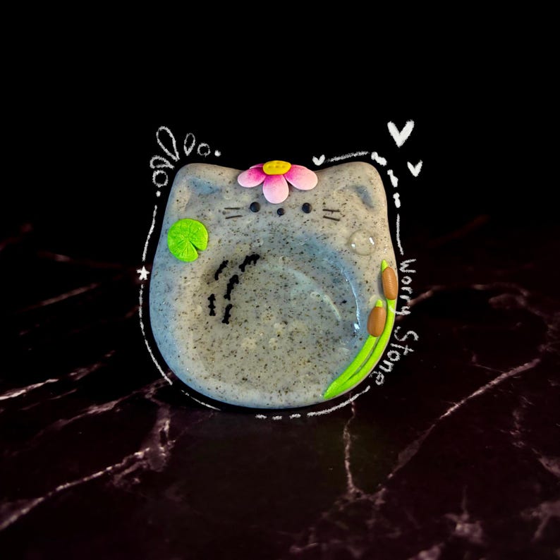 May include: A speckled gray cat-shaped worry stone featuring a pink flower on its head, a green lily pad, and brown cattails. The surface includes small black bird silhouettes and the text Worry Stone written in white script along the side.