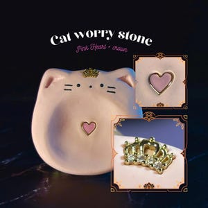May include: A light pink cat-shaped worry stone featuring a small gold crown with clear stones on the forehead and a pink heart inlay in the center. The text "Cat worry stone" and "Pink Heart + crown" is printed above the object in white and pink.