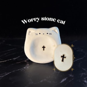May include: A white ceramic worry stone cat with a small black cross in the center, featuring simple black line details for eyes and whiskers. The text "Worry stone cat" is printed above, and a small oval inset shows a close-up of the cross.