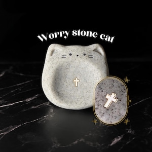 May include: A light gray speckled stone worry stone cat with a smooth, rounded shape and a small gold-colored cross inlay in the center. The cat features simple black line details for eyes and whiskers. Text above reads Worry stone cat.
