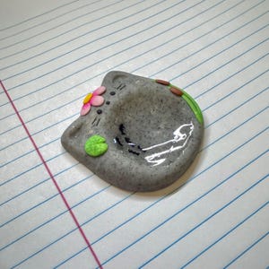 May include: A gray speckled cat-shaped trinket dish with a glossy finish. The dish features a small pink and yellow flower on the left ear, a bright green lily pad on the lower left, and a green stem with brown buds along the upper right edge.