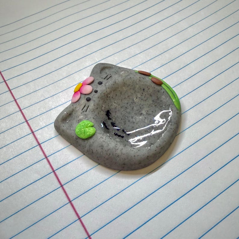 May include: A gray speckled cat-shaped trinket dish with a glossy finish. The dish features a small pink and yellow flower on the left ear, a bright green lily pad on the lower left, and a green stem with brown buds along the upper right edge.