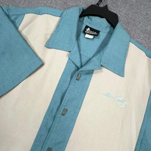 May include: A short-sleeve button-down shirt featuring a color-blocked design with teal panels and a cream-colored center. The shirt includes a collar, a black brand tag at the neck, and the embroidered text Elvis Presley in light blue on the chest.