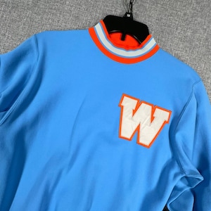 May include: A light blue long-sleeve sweatshirt featuring a mock neck collar with orange, white, and light blue stripes. A large white letter W with an orange border is appliqued on the front chest. The garment is displayed on a black hanger.