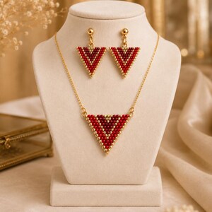 May include: A jewelry set consisting of a necklace and matching drop earrings. Each piece features a triangular pendant crafted from small red and gold beads arranged in a chevron pattern, suspended from a thin gold-tone metal chain.