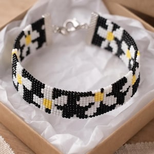 Puede incluir: Una pulsera de cuentas tejidas con un patrón repetitivo de flores de margarita blancas con centros amarillos sobre un fondo negro sólido. La pulsera está terminada con terminales de metal plateado y un cierre de mosquetón.