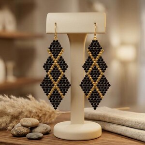 May include: A pair of long, geometric drop earrings featuring a repeating diamond pattern constructed from small, round black beads accented with gold-toned beads. The earrings hang from gold-toned metal hooks on a cream-colored T-shaped display stand.