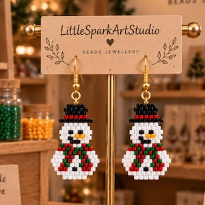 May include: A pair of beaded snowman earrings featuring a white body, a black top hat, and a red and green striped scarf. The earrings are suspended from gold-toned hooks and displayed on a rectangular cardboard card labeled LittleSparkArtStudio.