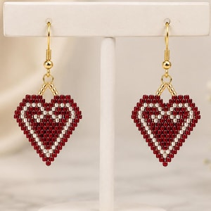 May include: A pair of heart-shaped dangle earrings crafted from small red and white beads. The earrings feature a gold-toned metal hook and a geometric heart pattern with a solid red center surrounded by a white beaded border and gold accents.