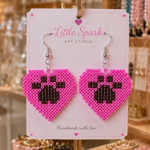 May include: A pair of heart-shaped earrings made from pink and black plastic beads, featuring a black paw print design in the center. The earrings are attached to a white display card labeled Little Spark Art Studio and Handmade with love.